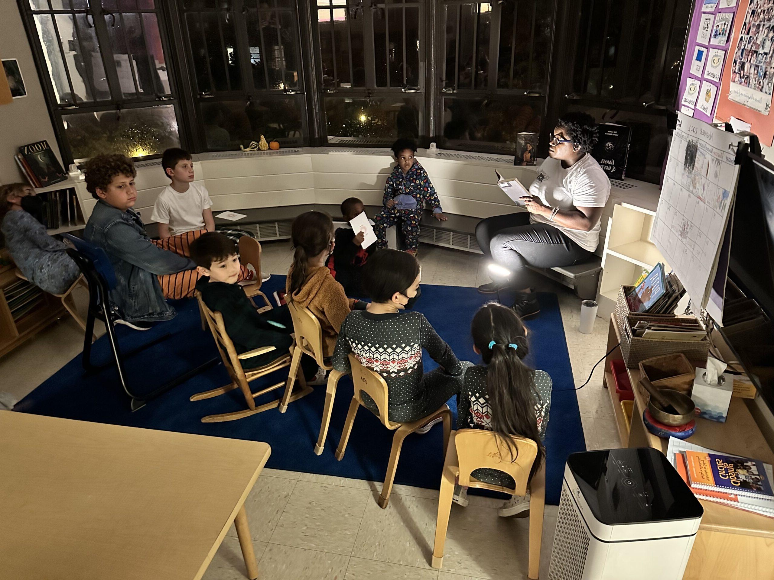 Fieldston Lower Assistant Principal Naomi Randolph reads to a room full of students in the dark; they sit on the floor in pajamas and listen along.